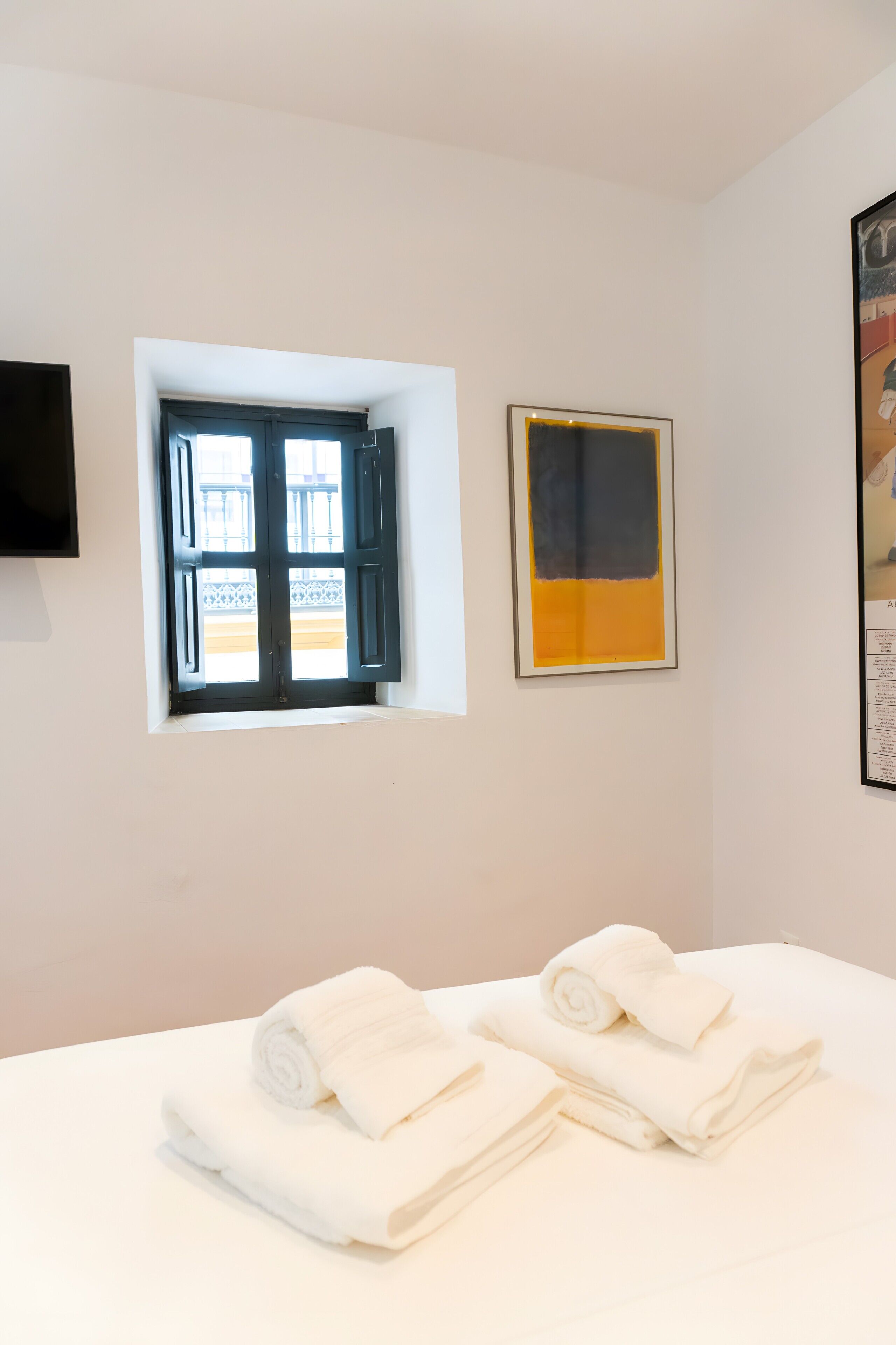 A hotel room with a bed, a window with open shutters, a framed artwork, and a flat-screen TV mounted on the wall.