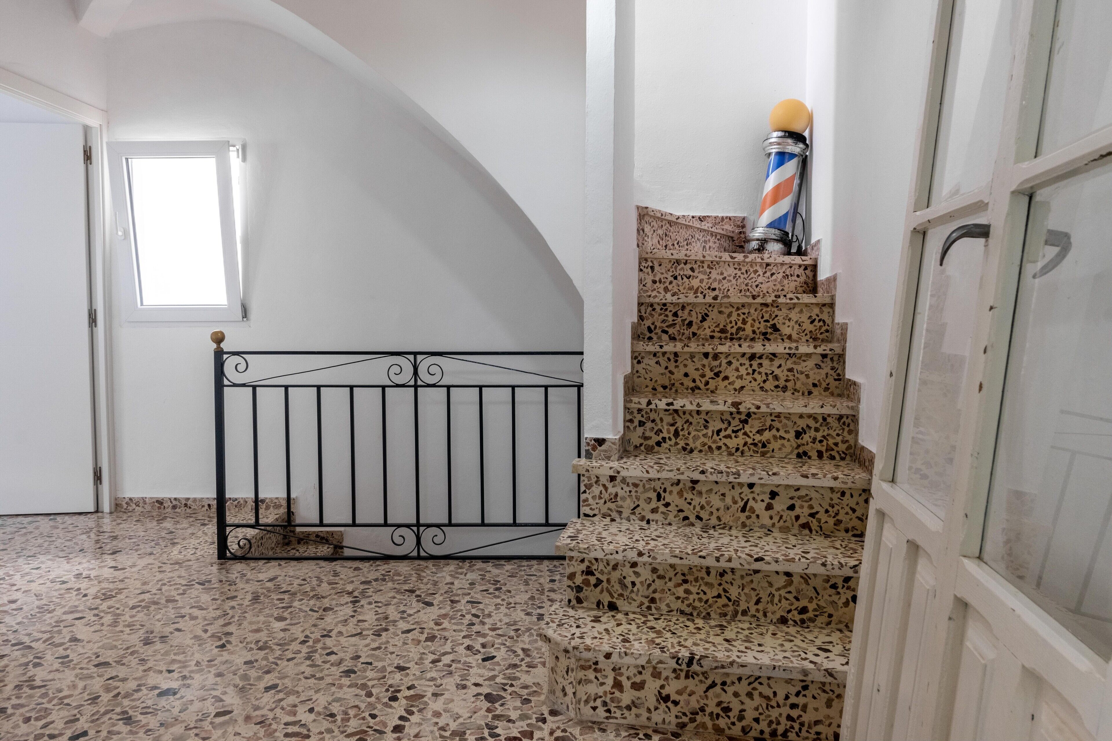 Una escalera con piso de terrazo que lleva a un rellano, una barandilla de metal y un palo de barbero.