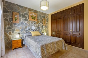 A bedroom with a stone wall, a bed with a patterned bedspread, a nightstand, a mirror, and a wooden wardrobe. - Holiday Home 'Echizzo' with Private Pool, Wi-Fi and Air Conditioning (Chiclana de la Frontera)