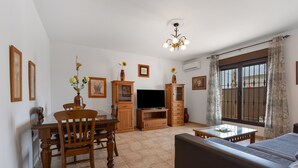 Ein Wohnzimmer mit Esstisch, Stühlen, einer Couch, einem Fernseher und einem Fenster mit Vorhängen.