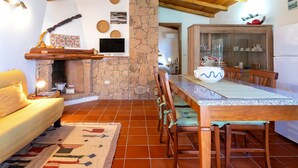Un salon chaleureux avec une cheminée en pierre, des poutres apparentes en bois au plafond, une table à manger avec des chaises et un buffet aux portes vitrées.