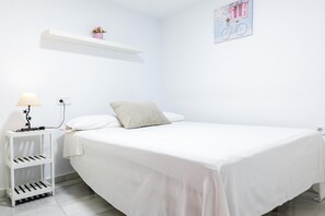 A white room with a bed, a nightstand with a lamp, a shelf with flowers, and a wall decoration.