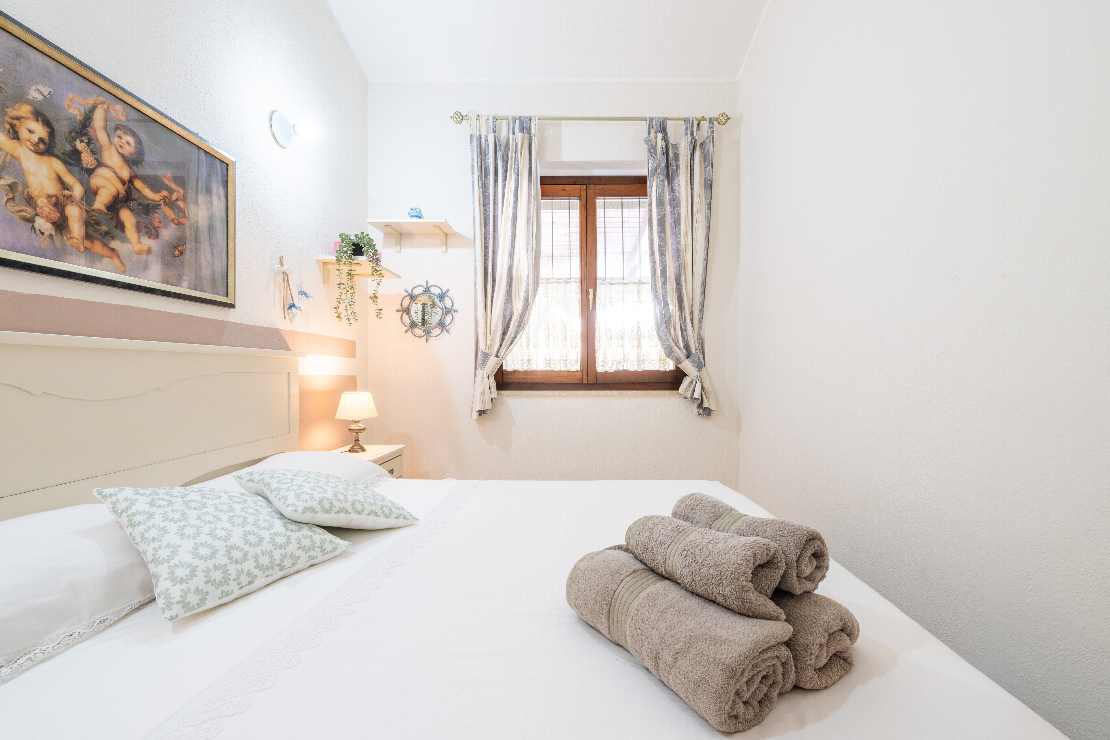 A hotel room with a bed, pillows, a painting, a window with curtains, and a bedside lamp.