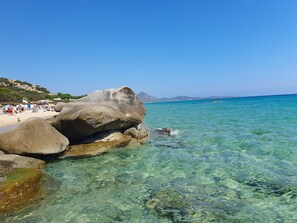 Une plage à l’eau claire, bordée de gros rochers, d’où l’on aperçoit des montagnes au loin.