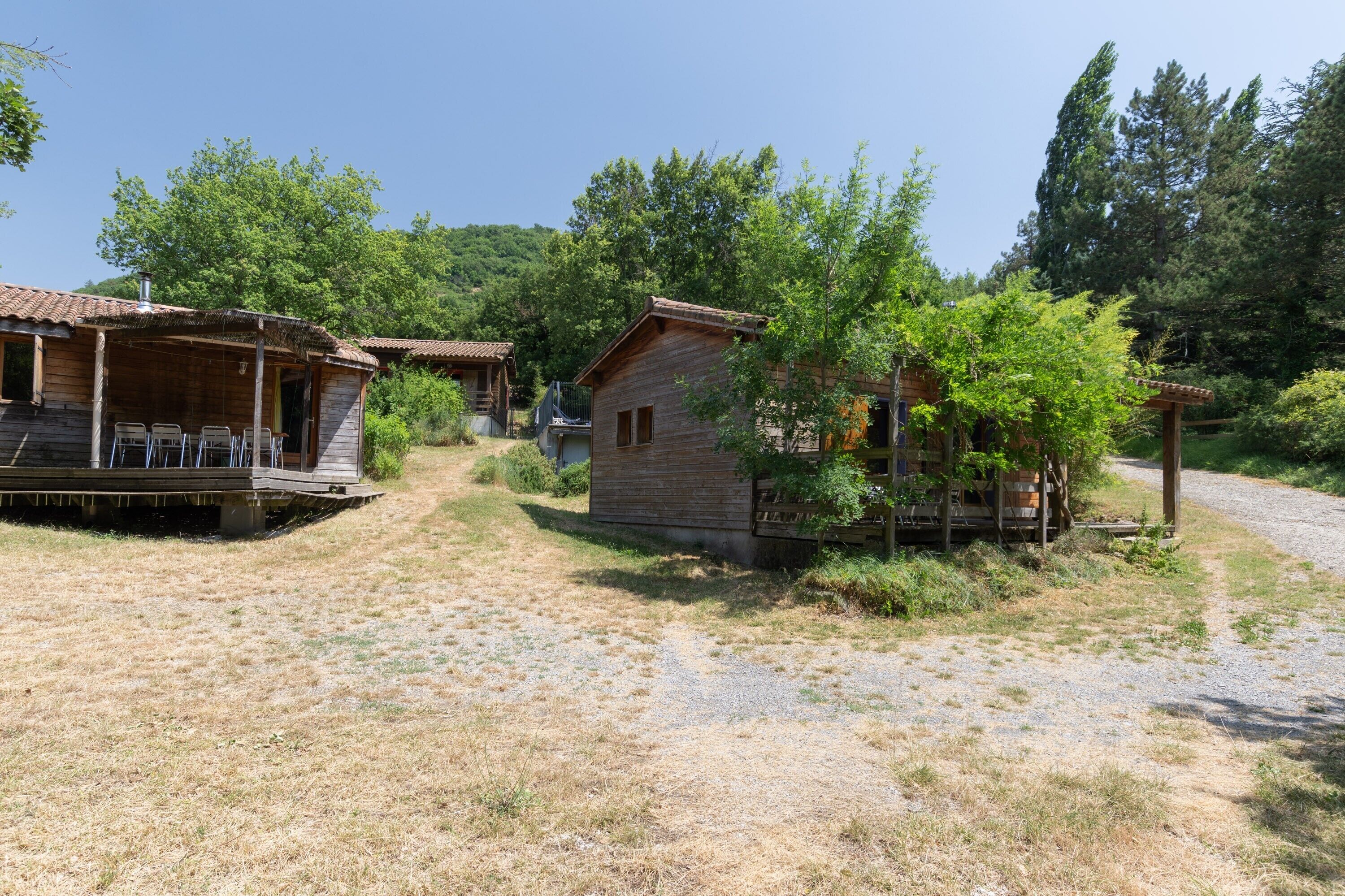 Une cabane en bois rustique, dotée d’un porche et de meubles d’extérieur, entourée d’arbres et bordée d’un chemin de gravier.