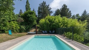 Ein rechteckiger Swimmingpool mit Holzterrasse, umgeben von üppiger Bepflanzung und einem Metallzaun.
