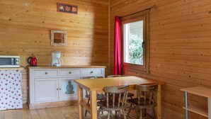 Interior de uma cabana de madeira com cozinha, mesa de jantar e janela com cortina vermelha.