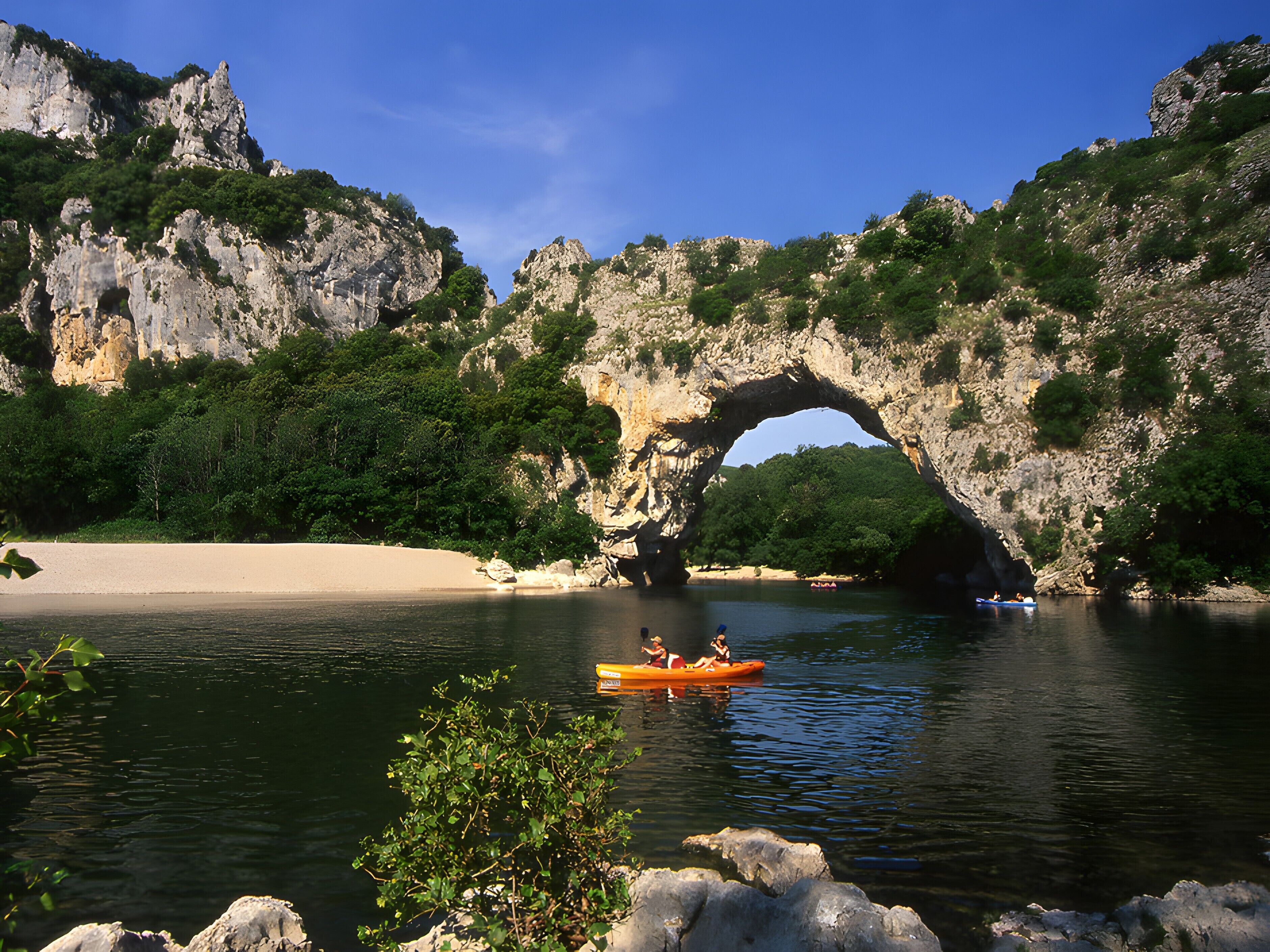 Une rivière traversée par un arc rocheux naturel, bordée de verdure et d’une plage de sable.