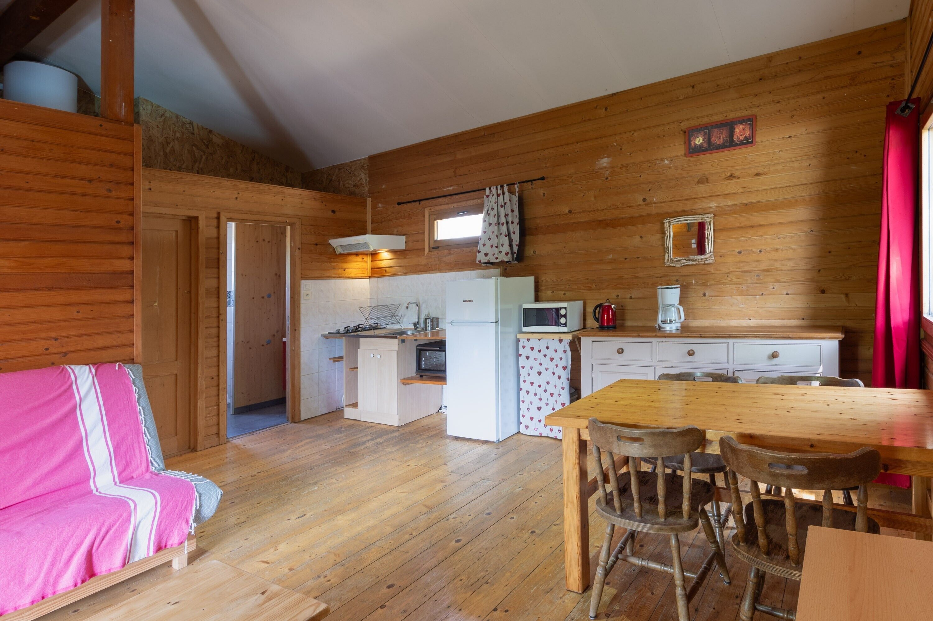 A cozy cabin interior with a wooden floor, a dining table with chairs, a kitchen area with appliances, and a pink sofa.