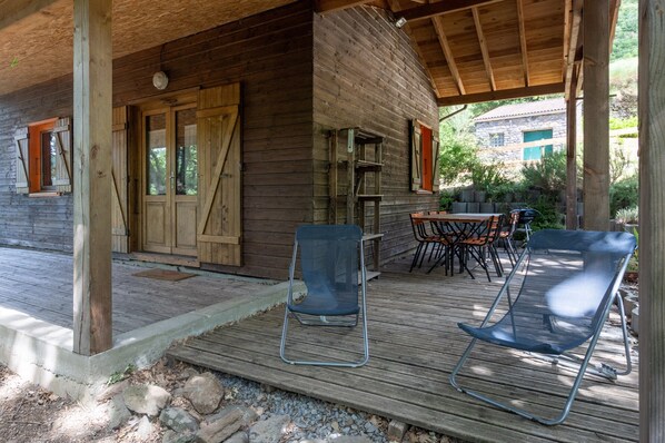 Une cabane en bois avec une véranda, des sièges d’extérieur et un bâtiment en pierre en arrière-plan.