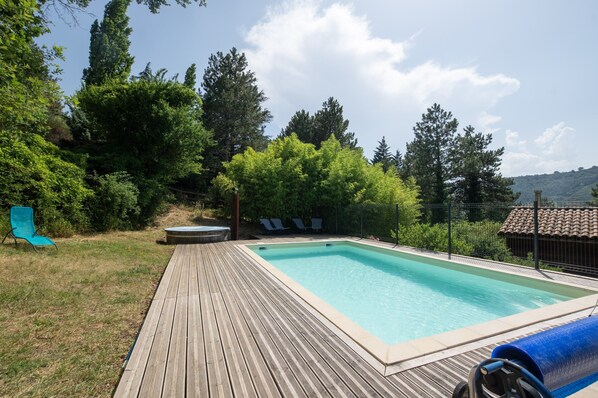 A rectangular swimming pool with a wooden deck, surrounded by greenery and trees.