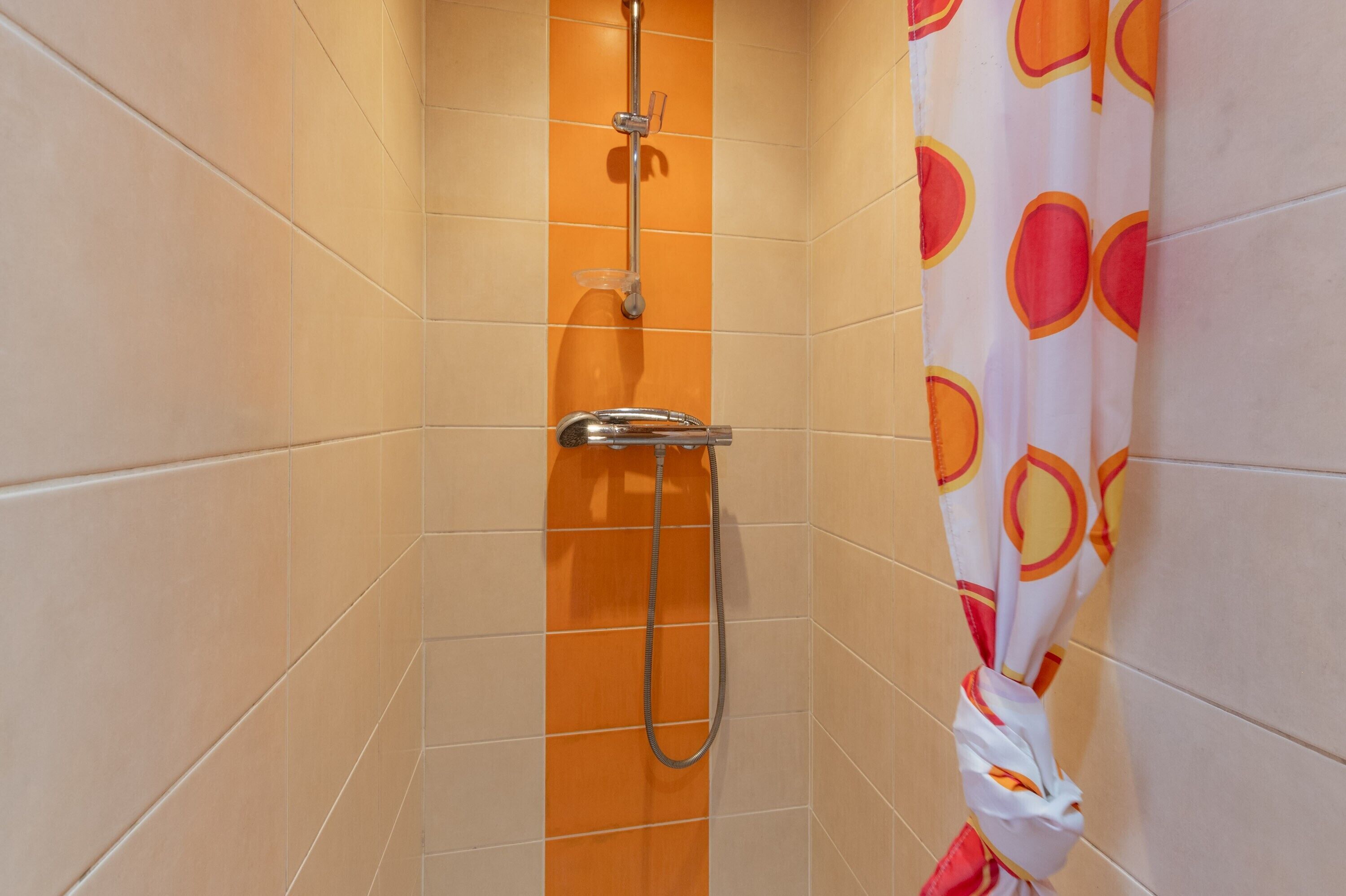 A shower with a handheld showerhead and a wall-mounted faucet, orange-tiled walls, and a shower curtain with a fruit pattern.