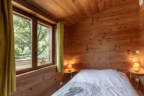 A wooden cabin room with a bed, two bedside lamps, a window with curtains, and a view of trees.