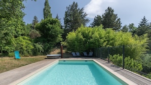 Una piscina rectangular con un muelle de madera, rodeada por una valla metálica y árboles.