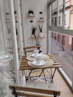 A modern interior with a wooden table set for two, a window with a view of the street, and potted plants on shelves.