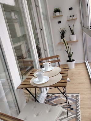 Ein Holztisch mit weißen Tellern und Tassen, eine Bank und ein gemustertes Teppich auf einem Balkon mit Blumentöpfen und Blick ins Innere.