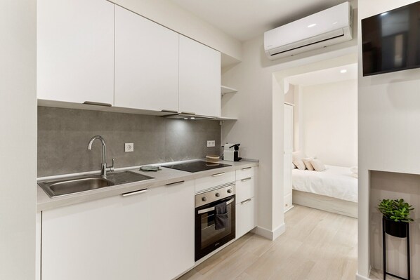 A modern kitchen with white cabinets, a stainless steel sink, a built-in oven, and an air conditioning unit above the counter.