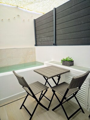 A small outdoor seating area with two wicker chairs and a table, a small plant on a shelf, and a glass partition separating the seating from a pool area.