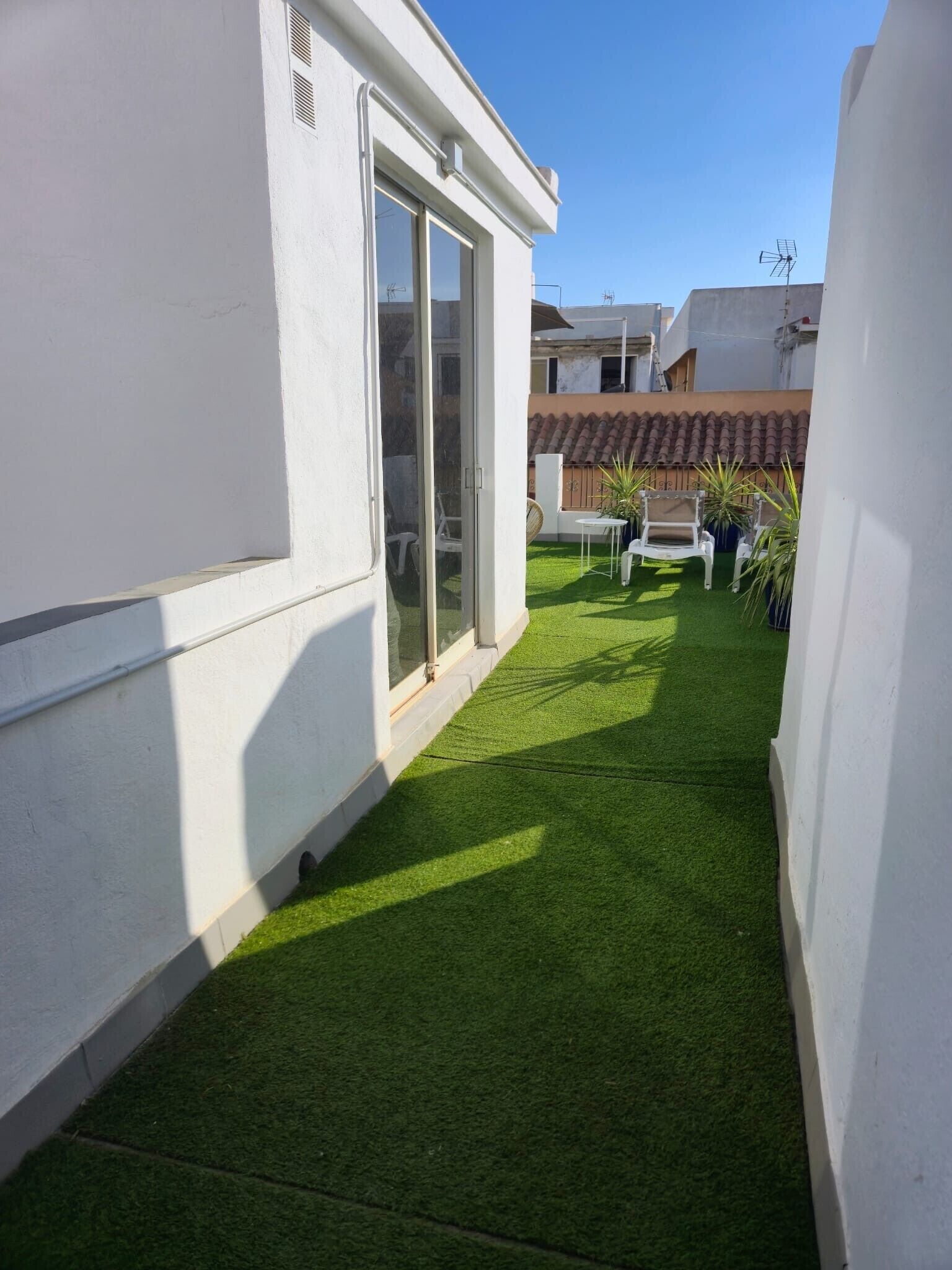 Un patio estrecho con césped artificial verde, una puerta corrediza de vidrio y mobiliario exterior blanco.