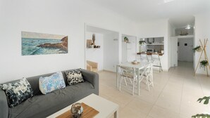 A modern living room with a grey sofa, a dining table with chairs, a kitchen area, and a painting on the wall.