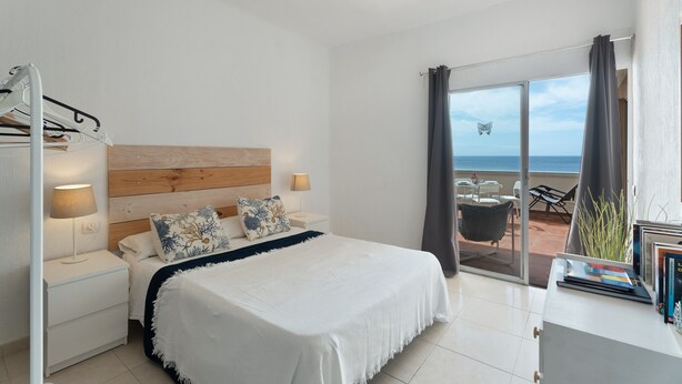 A bedroom with a bed, nightstands, a lamp, a window with curtains, and a view of the sea.