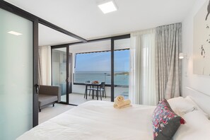 A hotel room with a bed, a chair, a table, and a view of the sea.