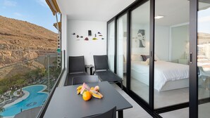Un balcone con vista sulla piscina, un tavolo da pranzo con frutta, due sedie e un letto con piumone bianco.