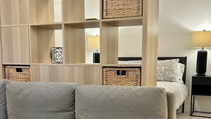 Un salon moderne avec un canapé gris, une étagère en bois agrémentée de paniers en osier, une table de chevet avec une lampe et un lit avec une literie à motifs.