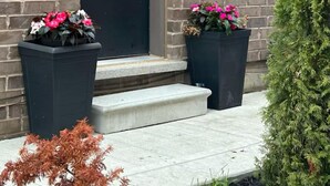 Un bâtiment en briques avec une porte sombre, deux grands jardinières fleuries et un petit arbre à feuilles persistantes.