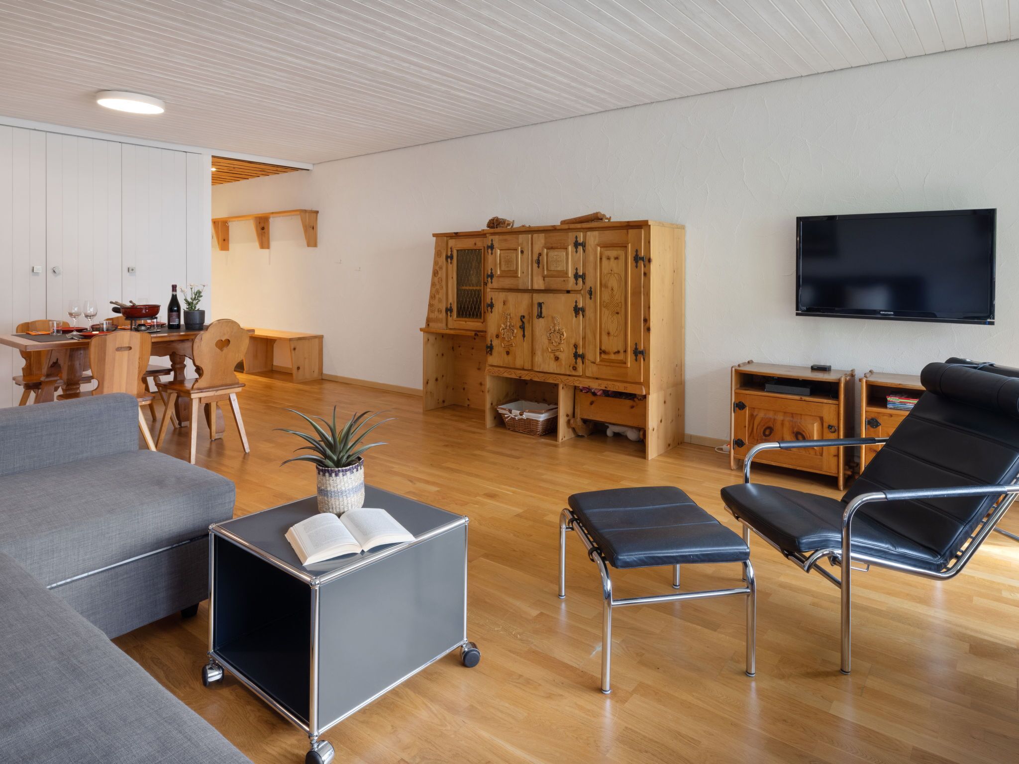 Un salon moderne avec un canapé gris, un fauteuil en cuir noir, un meuble en bois, un téléviseur et un coin repas avec des meubles en bois.