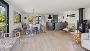 A spacious living area with a dining table, a sofa, a fireplace, and a kitchen in the background. - Cottage with fjord view in lovely Vellerup (Skibby)