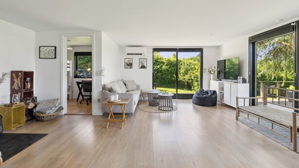 A spacious living area with a sofa, coffee table, and a TV. There is a large window with a view of greenery. - Cottage with fjord view in lovely Vellerup (Skibby)