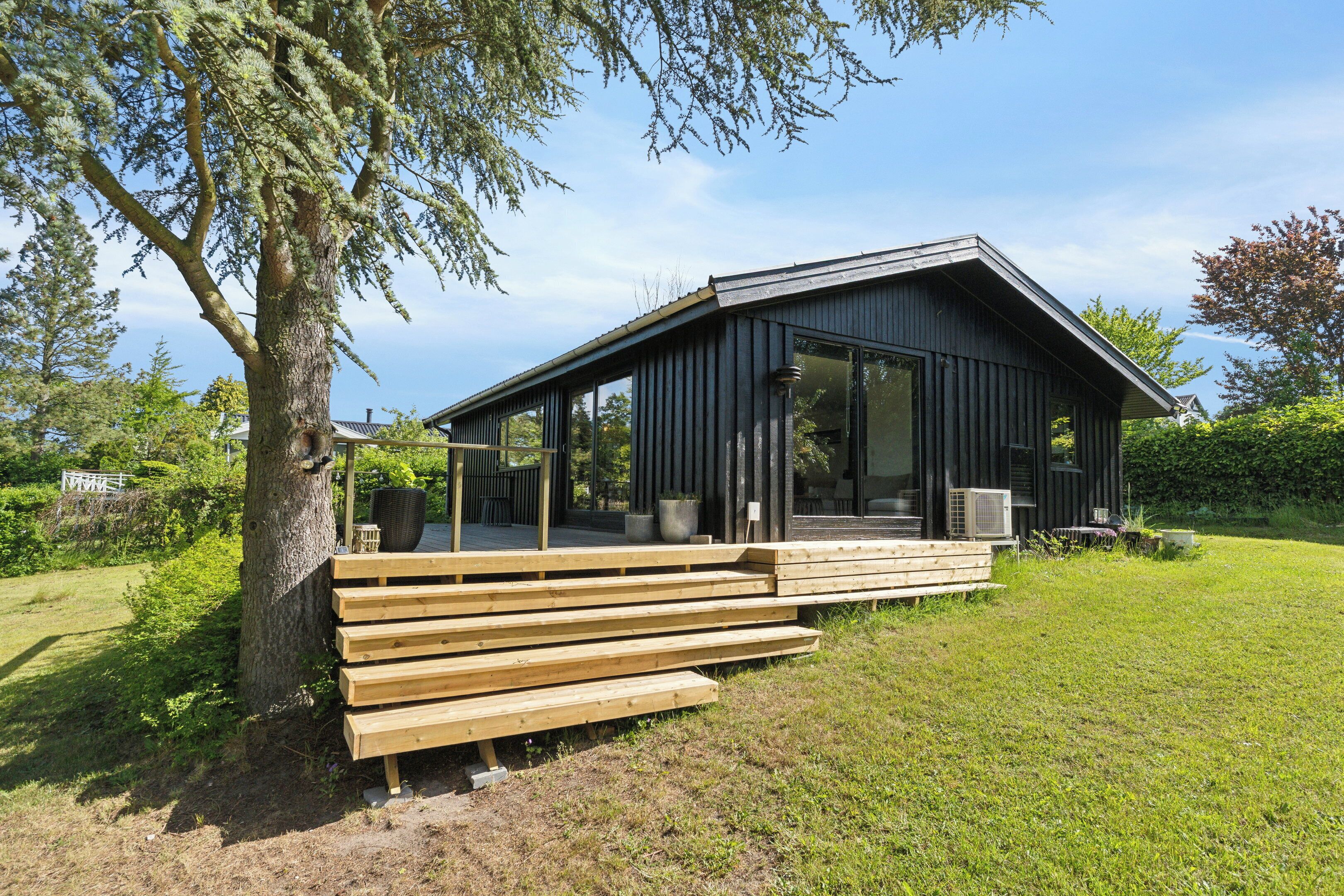 Una cabaña de madera moderna con terraza, ventanas grandes y un árbol cercano.