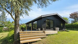 A modern wooden cabin with a deck, large windows, and a tree nearby. - Cottage with fjord view in lovely Vellerup (Skibby)