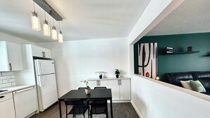 Une cuisine de taille réduite avec des meubles blancs, une table à manger noire et quatre chaises, ainsi que des luminaires suspendus.