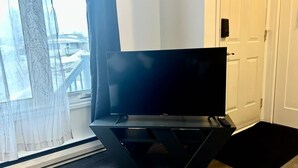 Un salon moderne avec un téléviseur à écran plat posé sur un meuble noir, de grandes fenêtres ornées de rideaux légers et une vue sur un balcon.