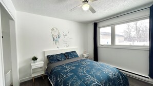 Une chambre à coucher comprenant un lit, une table de chevet, un ventilateur de plafond, une fenêtre et un motif décoratif sur le mur.