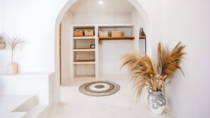 Un interior minimalista con un arco blanco que da a un espacio de almacenamiento con estanterías y cajas. Una alfombra tejida y un jarrón con plantas secas están sobre el suelo.