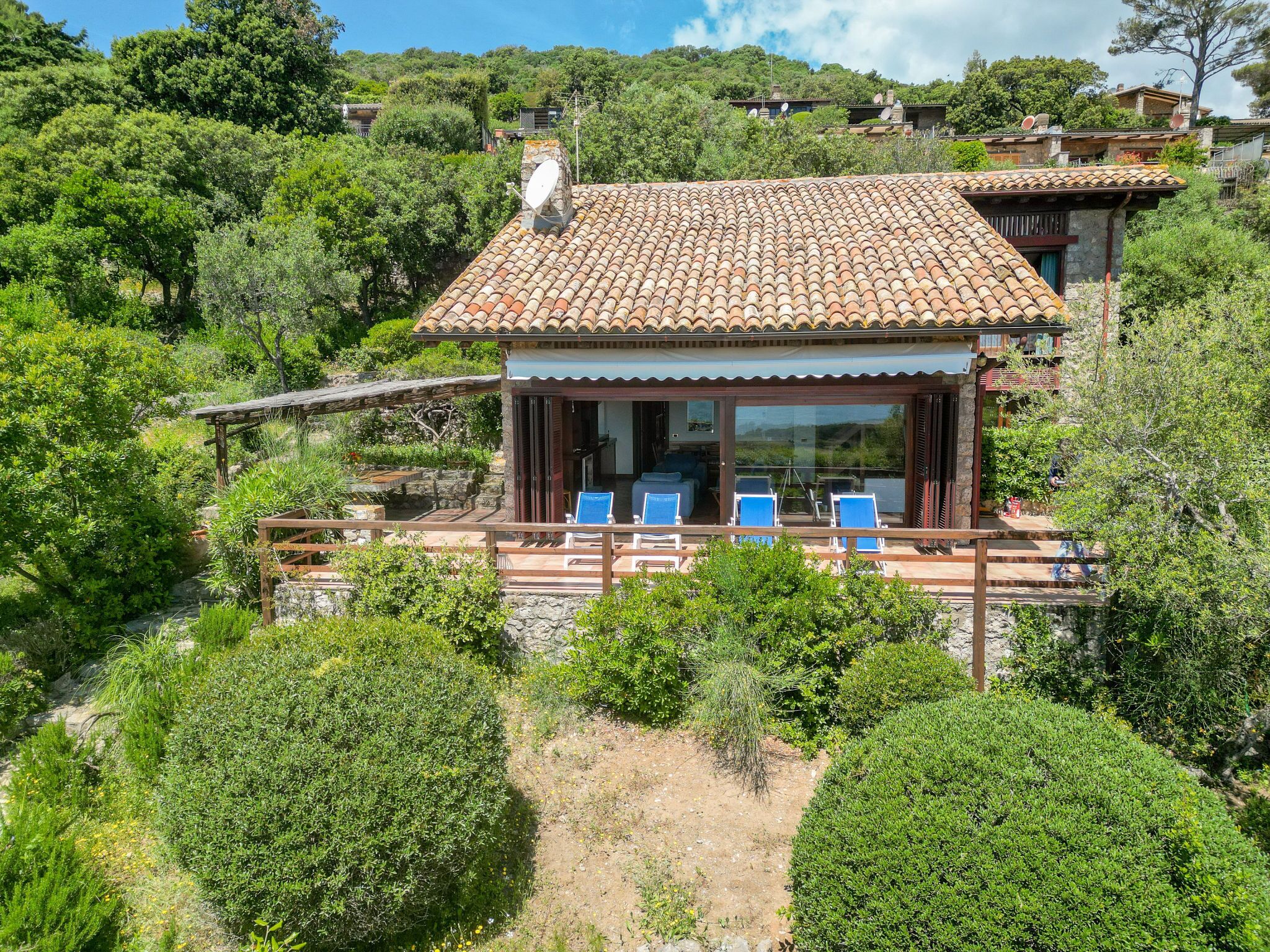 Eine Villa mit Ziegeldach, einer überdachten Terrasse mit blauen Stühlen und Blick auf die umgebende Grünfläche.