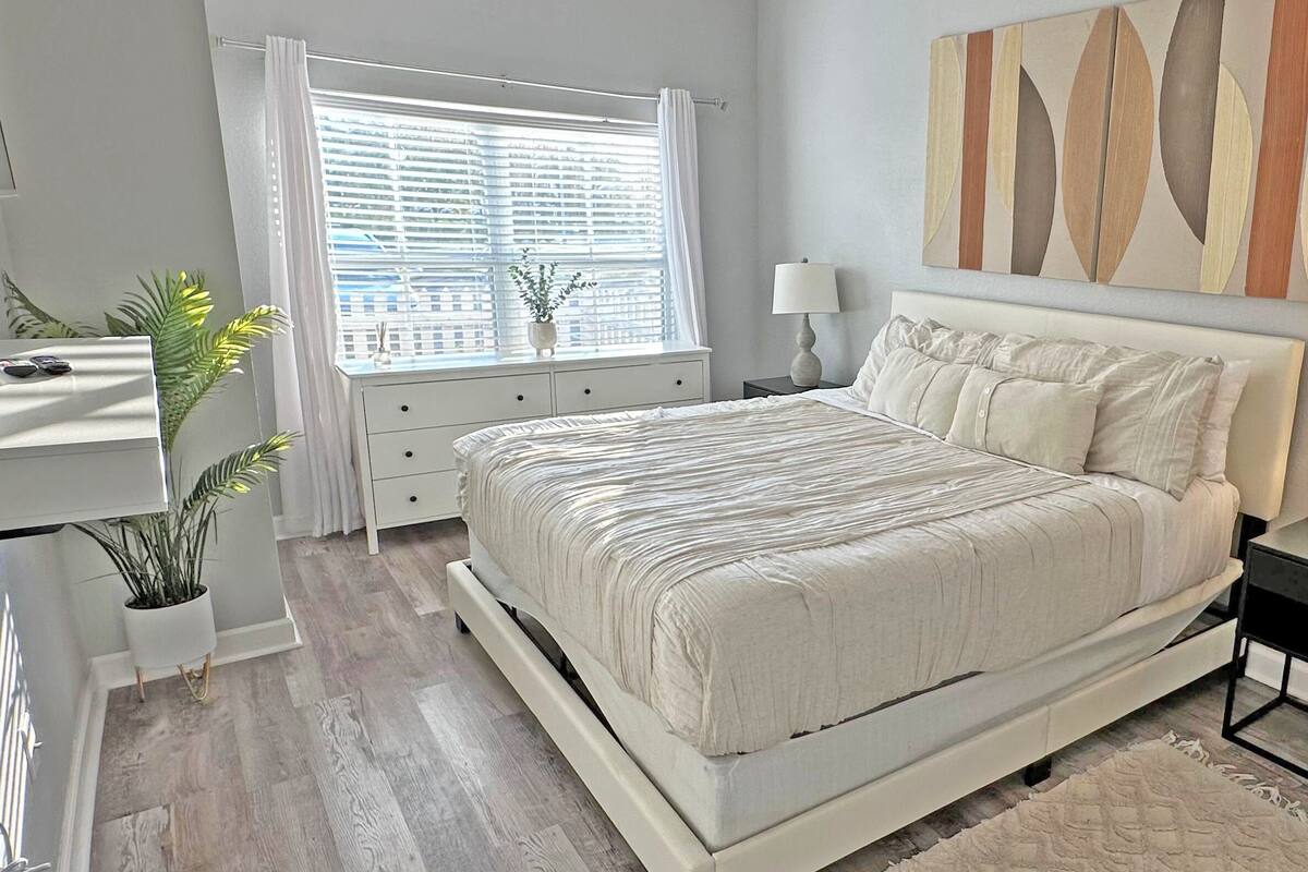 A modern bedroom with a large bed, a bedside table with a lamp, a dresser, and a window with blinds.