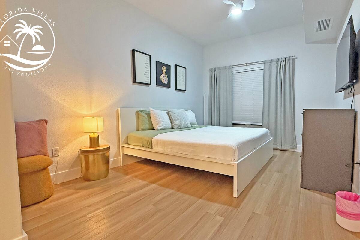 A bedroom with a bed, a nightstand, a lamp, a window with curtains, a TV mounted on the wall, and a trash bin.