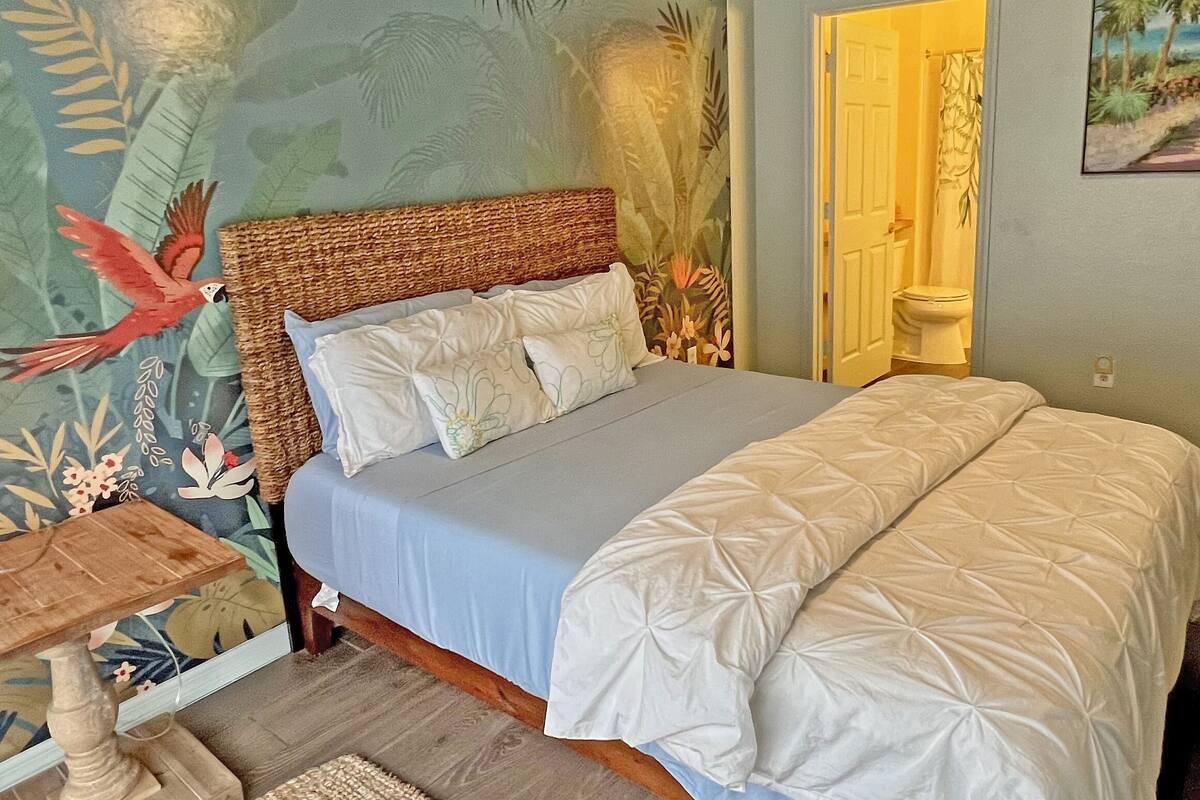 A bedroom with a bed, a nightstand, a wall mural, and a door leading to another room.
