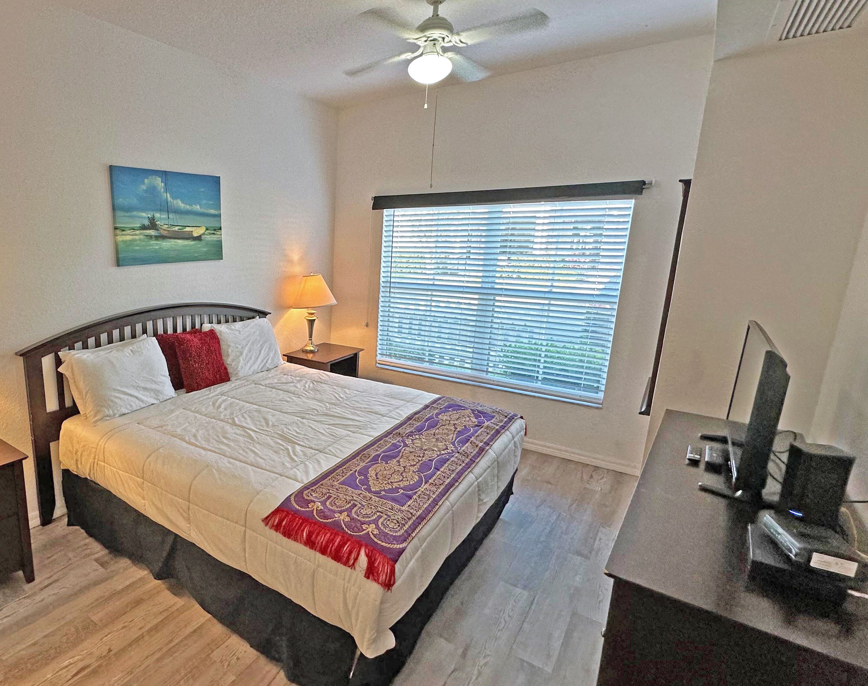 A bedroom with a bed, a nightstand, a lamp, a ceiling fan, a window with blinds, and a painting on the wall.