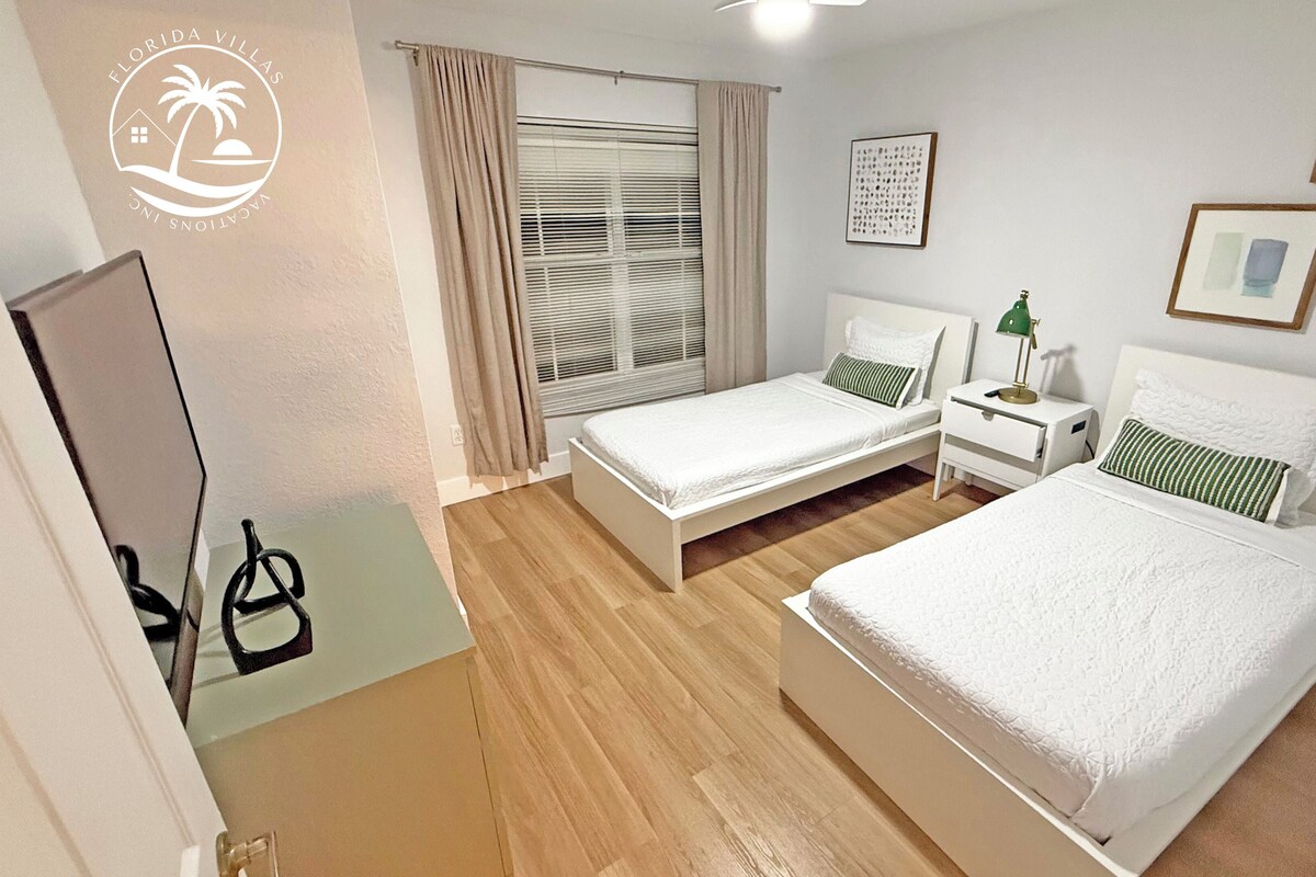 A hotel room with two beds, a window with blinds, a bedside table with a lamp, and a mirror.