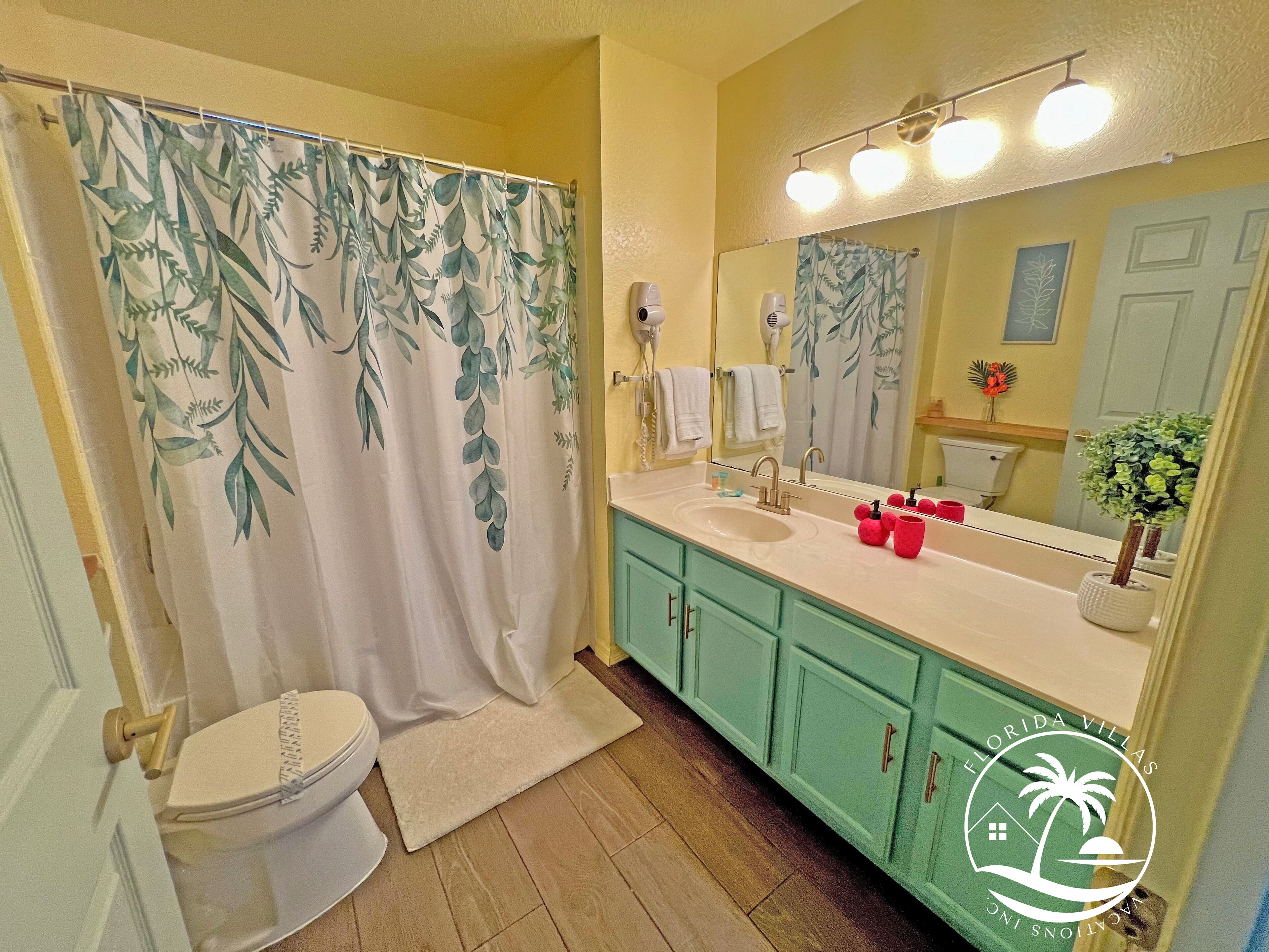 A bathroom with a toilet, a sink with a mirror, a shower curtain with a leaf pattern, and a vanity with a plant.