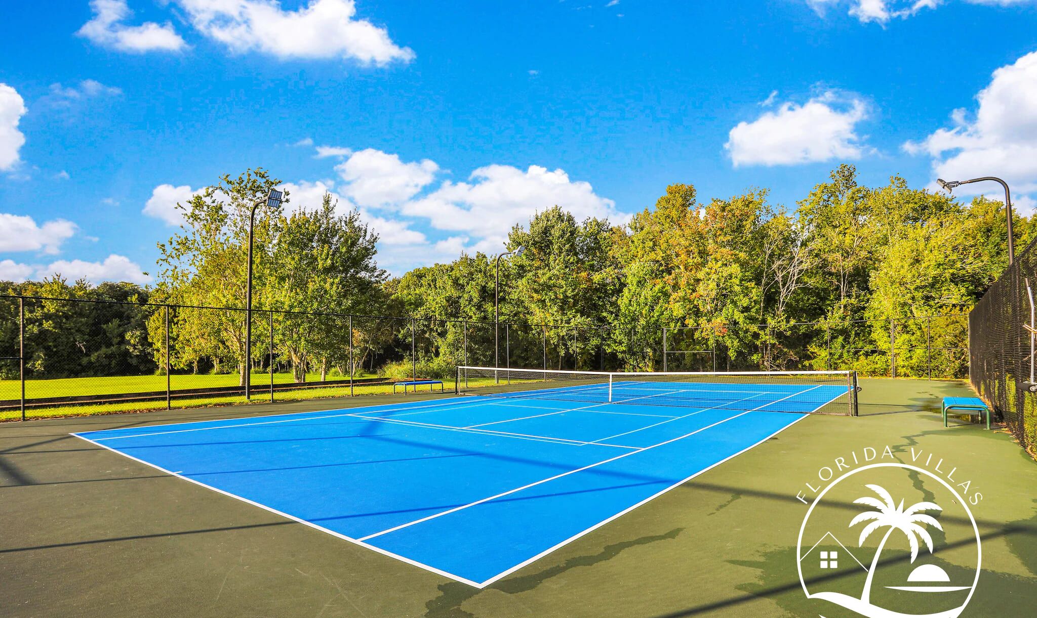 Un court de tennis extérieur doté d’une surface bleue, entouré d’arbres et d’une clôture.