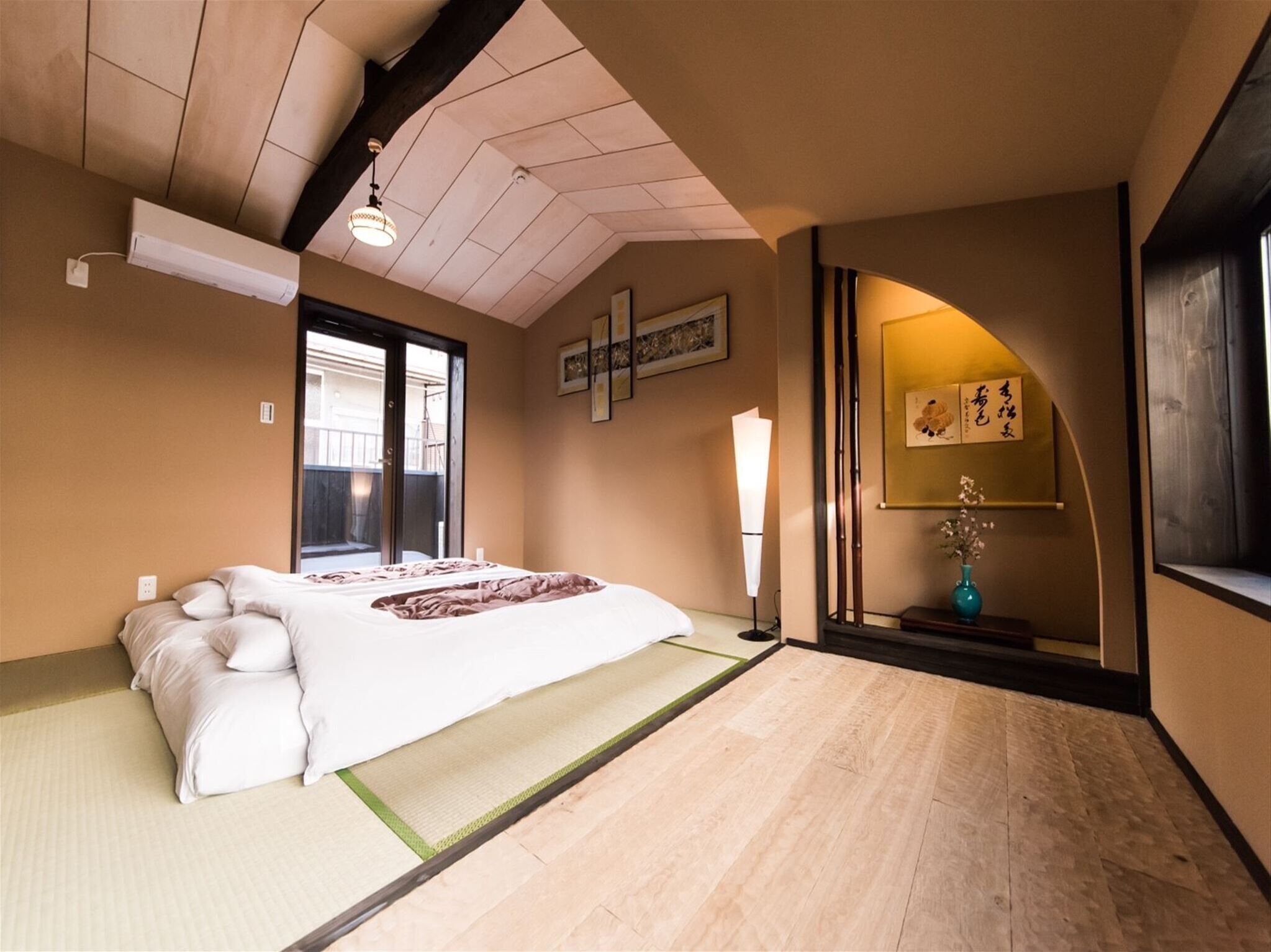 A traditional Japanese-style room with tatami flooring, a low bed with a white and pink bedding, a sliding door leading to an outdoor area, and a wall-mounted shelf with a vase and calligraphy.