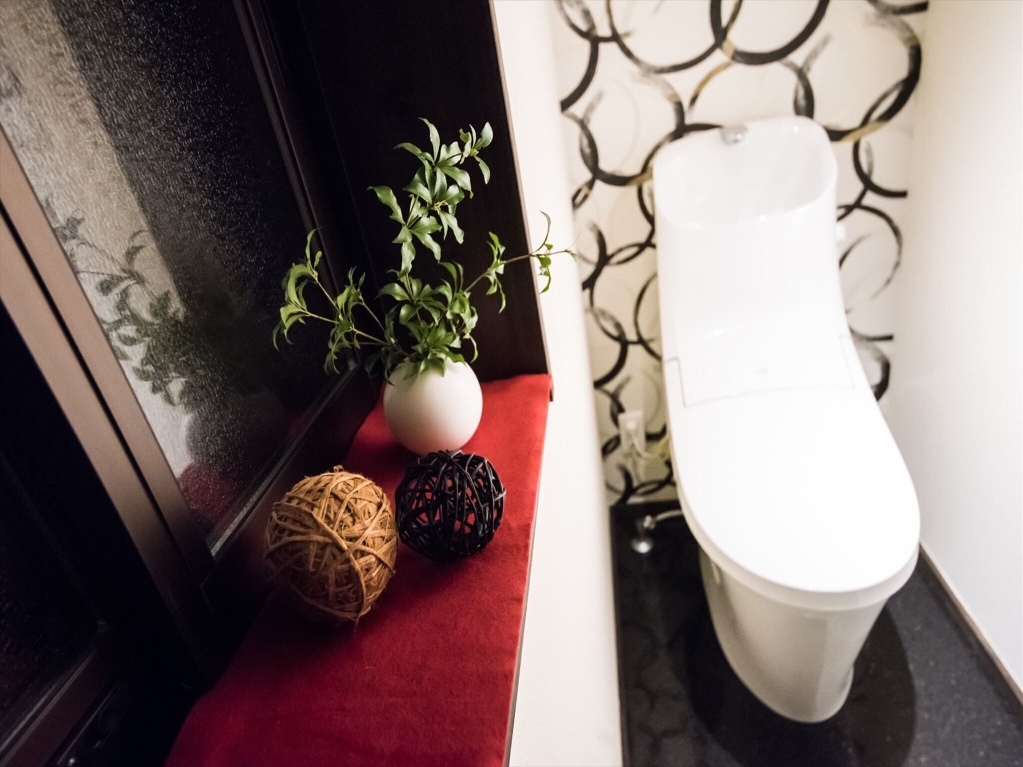 Un baño con un inodoro blanco, un banco rojo, un jarrón blanco con plantas verdes y dos adornos esféricos.