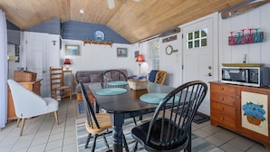 A cozy, well-lit dining area with a wooden ceiling, a dining table with chairs, a kitchen area with a microwave, and a wall-mounted shelf with decorative items.