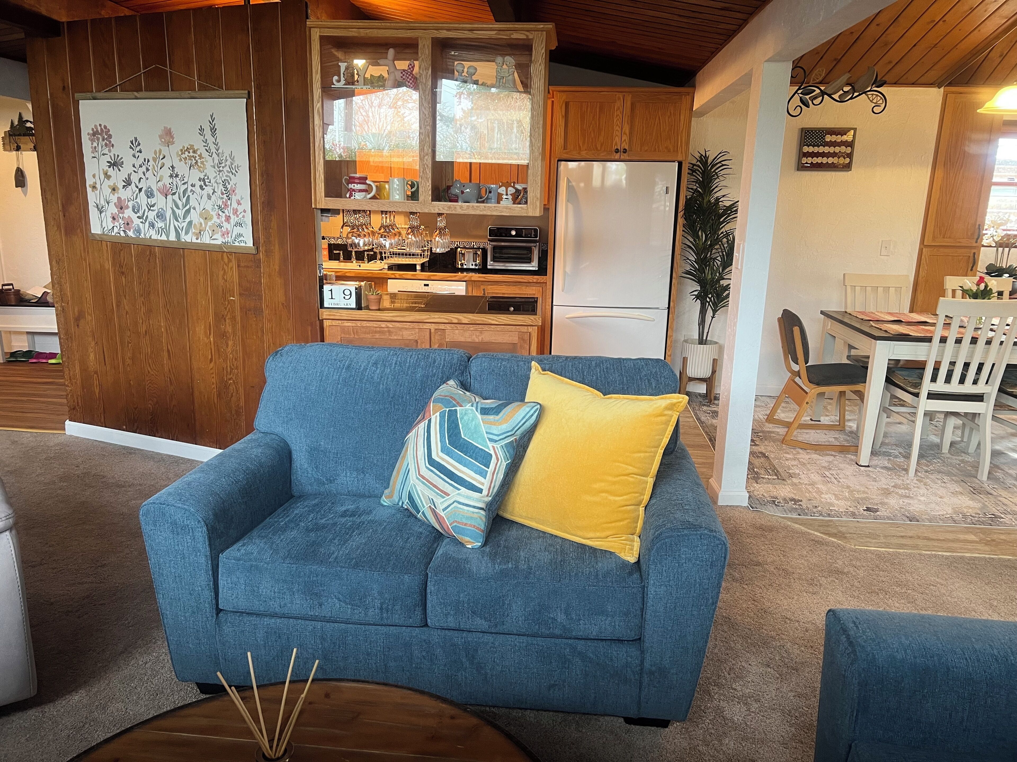 Un sofá azul con un cojín amarillo, un armario de madera con electrodomésticos de cocina y una zona de comedor con sillas blancas.